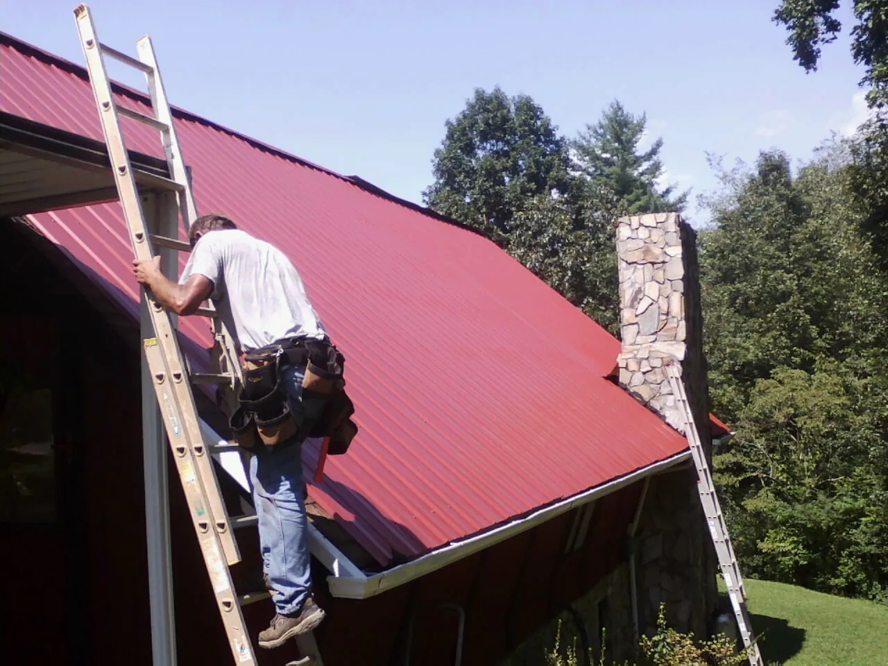 Roofer on a ladder beside a red metal roof and stone chimney — Soffit & Fascia Repair by Roofing Brothers in New Harmony